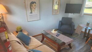 Un salón con un sofá beige, un cojín azul, una mesa de centro de cristal, un sillón gris, una mesita con una vela y una planta, y un cuadro en la pared.