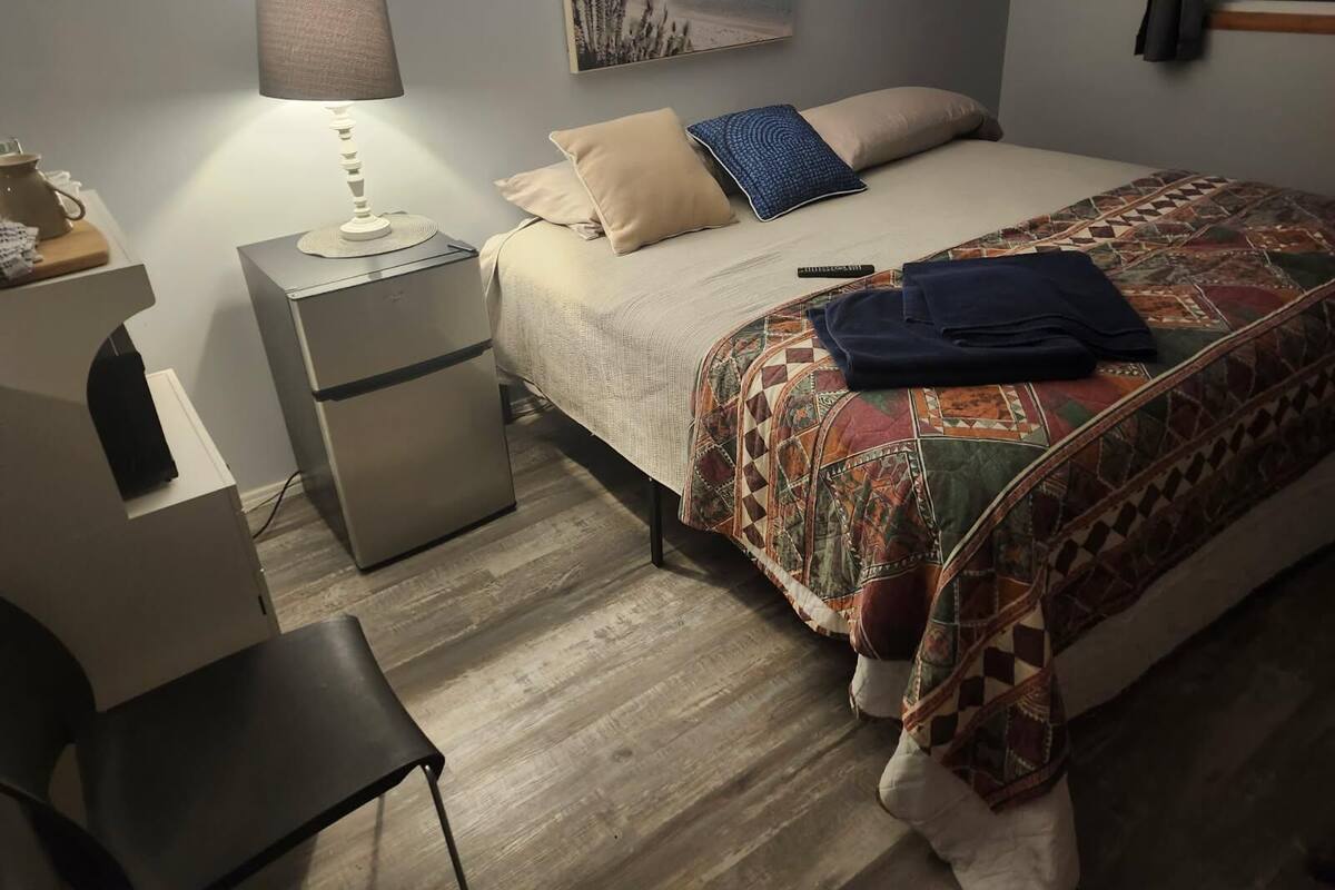 A bedroom with a bed, a nightstand, a lamp, a chair, and a small refrigerator.