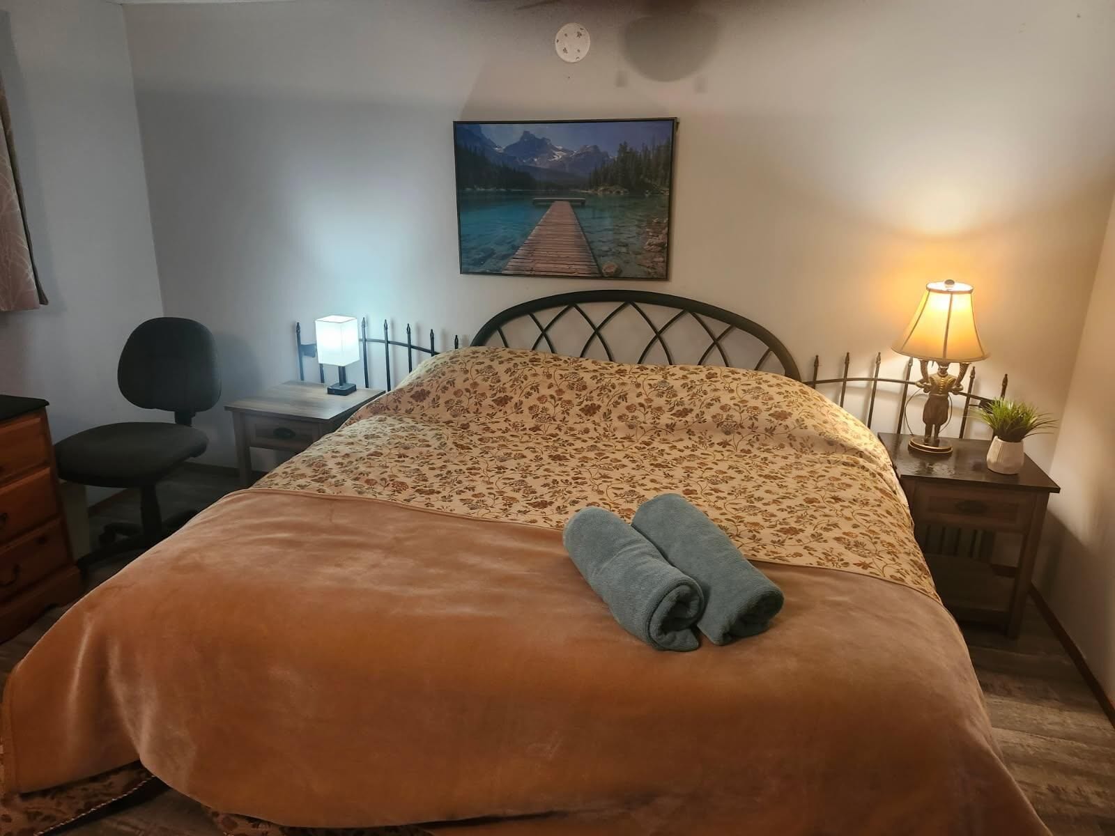 A neatly made bed with a floral bedspread, a rolled towel, a desk with a lamp, a chair, and a painting of a lake and mountains on the wall.