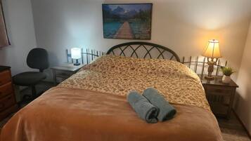 A neatly made bed with a floral bedspread, a rolled towel, a desk with a lamp, a chair, and a painting of a lake and mountains on the wall.