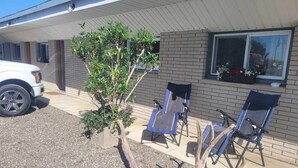 Un edificio con estacionamiento techado, un árbol pequeño y tres sillas de jardín azules.