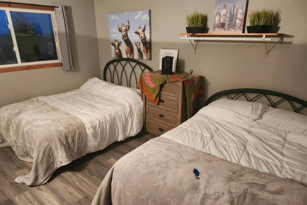 A bedroom with two single beds, a dresser, a window, and wall decorations including a painting of cows and a shelf with plants.