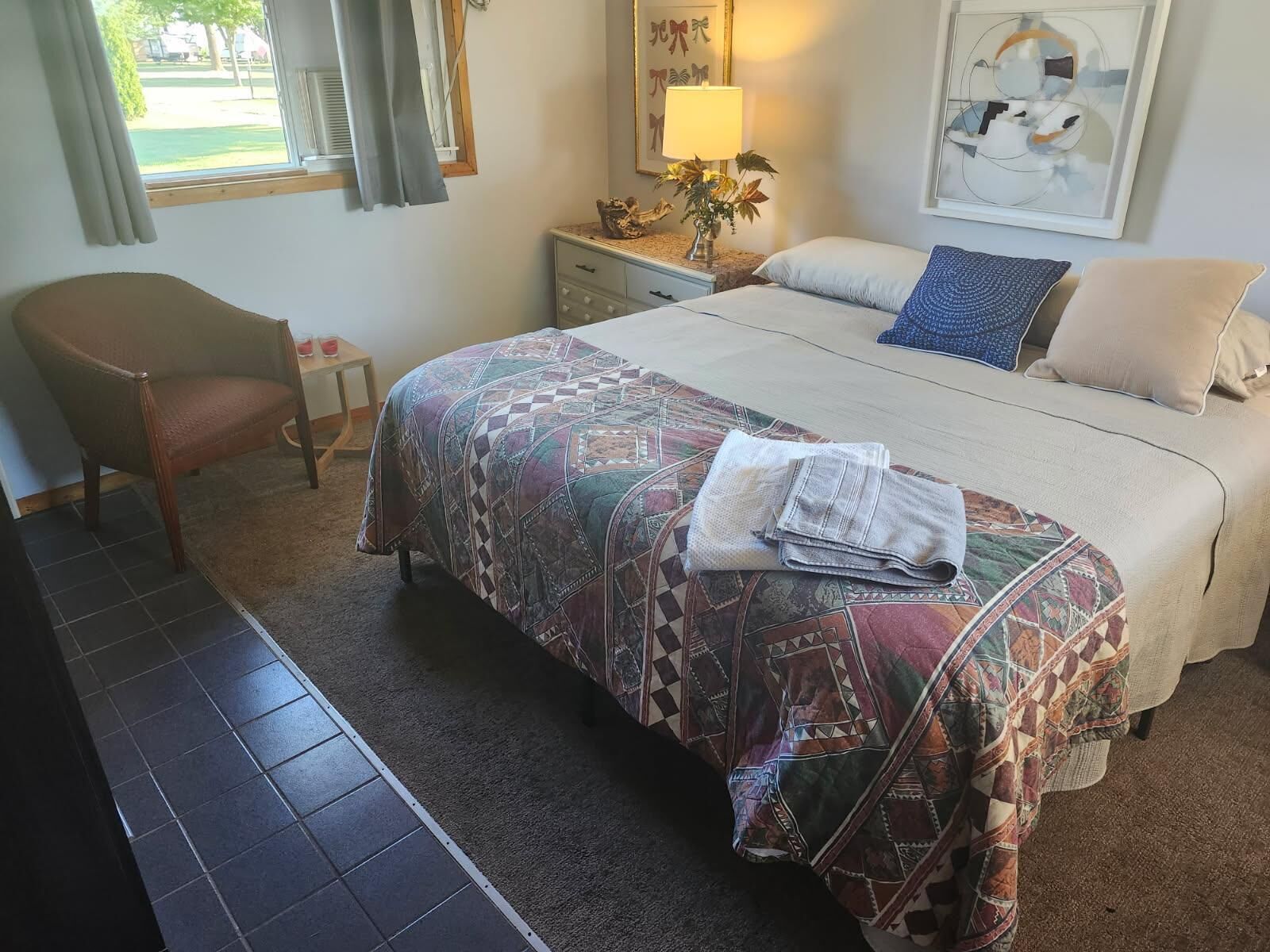 A bedroom with a bed, a chair, a nightstand, a lamp, and a window with curtains.