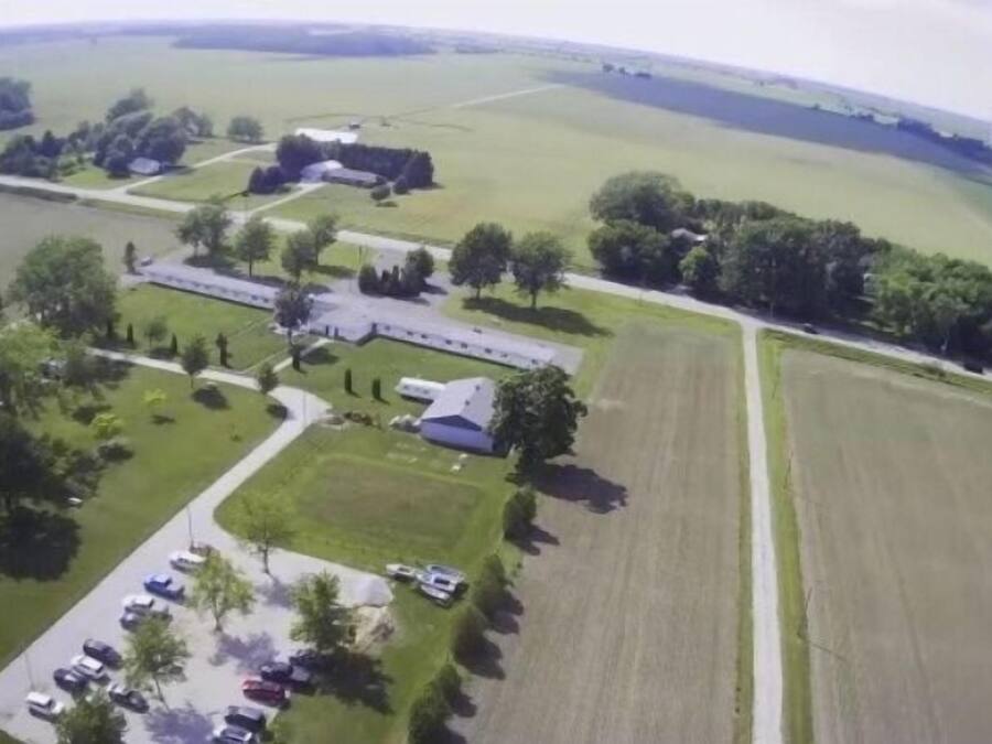 An aerial view of a rural property with a large field, a building with a sloped roof, and a parking area with several cars.