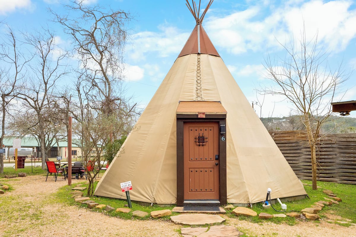 A teepee with a brown door, surrounded by a stone pathway, and a sign indicating a reservation requirement.