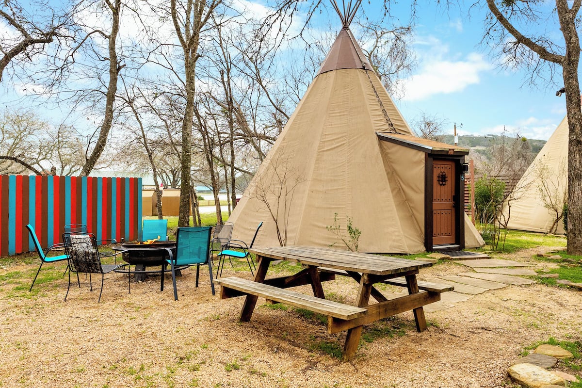A teepee-style tent with a wooden door, a picnic table, and chairs in a grassy area with trees.