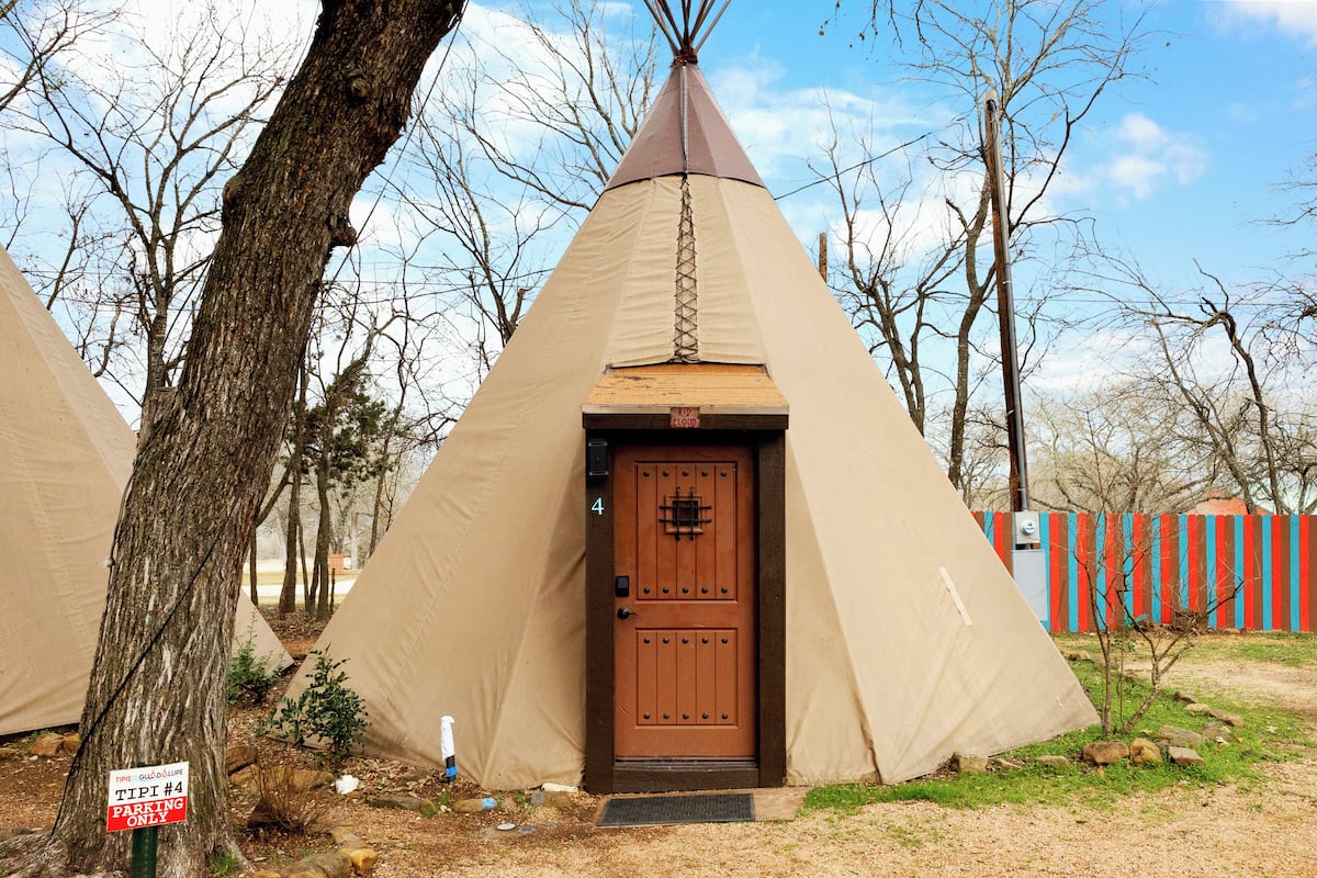 A teepee with a wooden door, surrounded by trees and a fenced area.