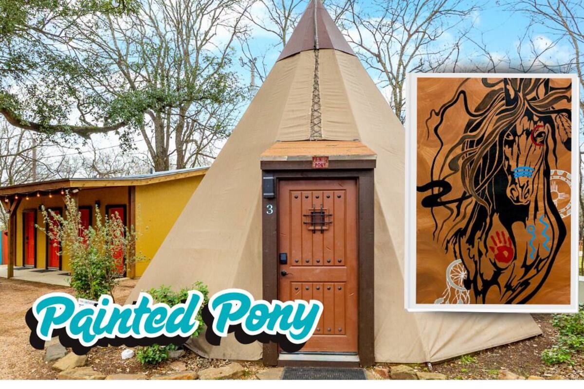 A painted pony-themed tent with a brown door, surrounded by trees and a building with red doors.