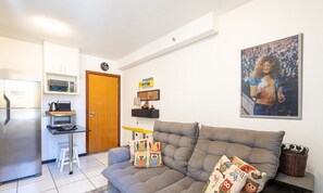 Una sala de estar moderna con un sofá gris, almohadones con forma de búho de colores, una zona de cocina con nevera, microondas y tostadora, y un póster enmarcado en la pared.