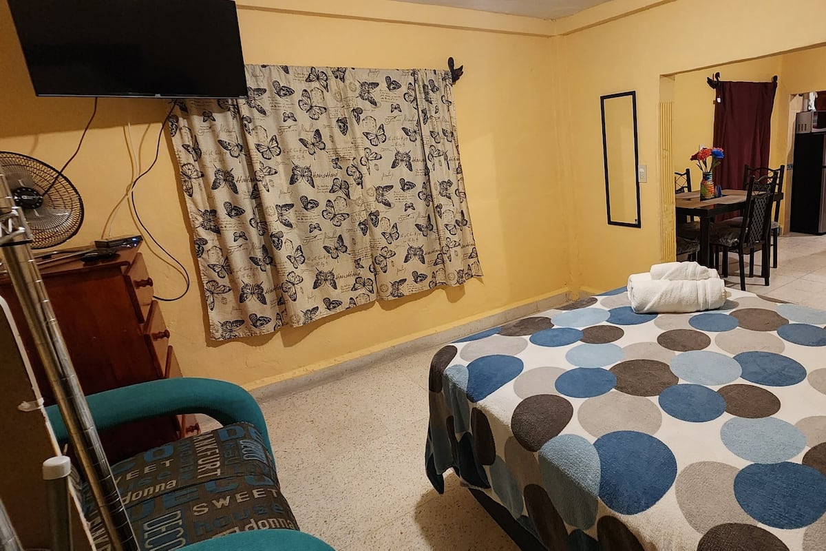 A bedroom with a bed, a desk, and a chair. There is a TV on the wall and a fan on the floor.
