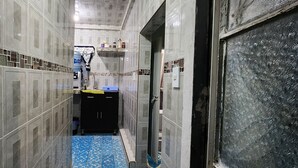 Un pasillo estrecho con paredes de azulejos y un suelo azul con estampado. Se observa un mueble negro con cajones y una estantería con diversos objetos.