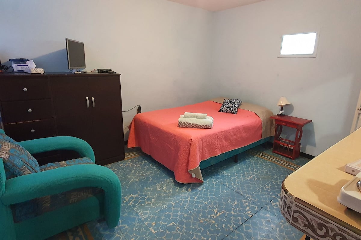 A small, compact room with a bed, a desk, a chair, a dresser, and a window.