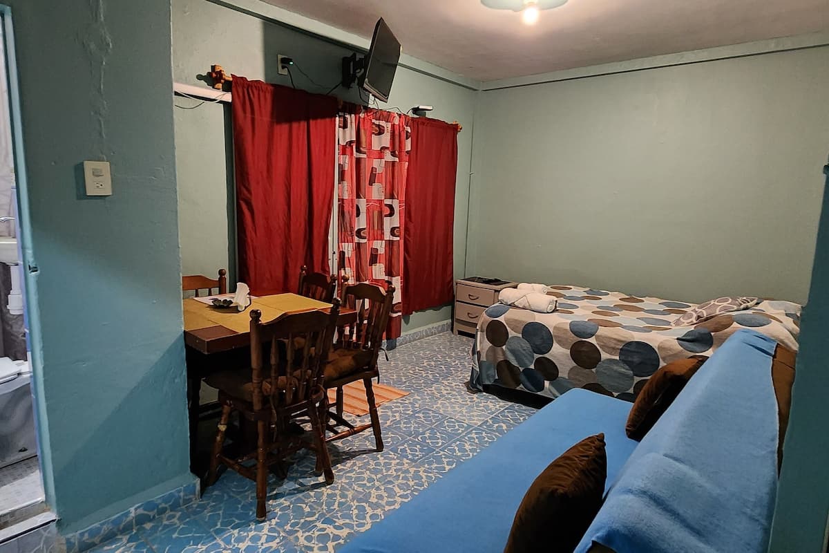 A small, compact room with a bathroom, a dining table with chairs, a bed with a patterned bedspread, and a television mounted on the wall.