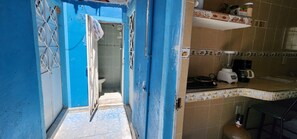 Ein schmales Gang führt zu einem Badezimmer mit Toilette und Dusche. Man sieht eine Küchenzeile mit Herd, Mixer und Spüle.