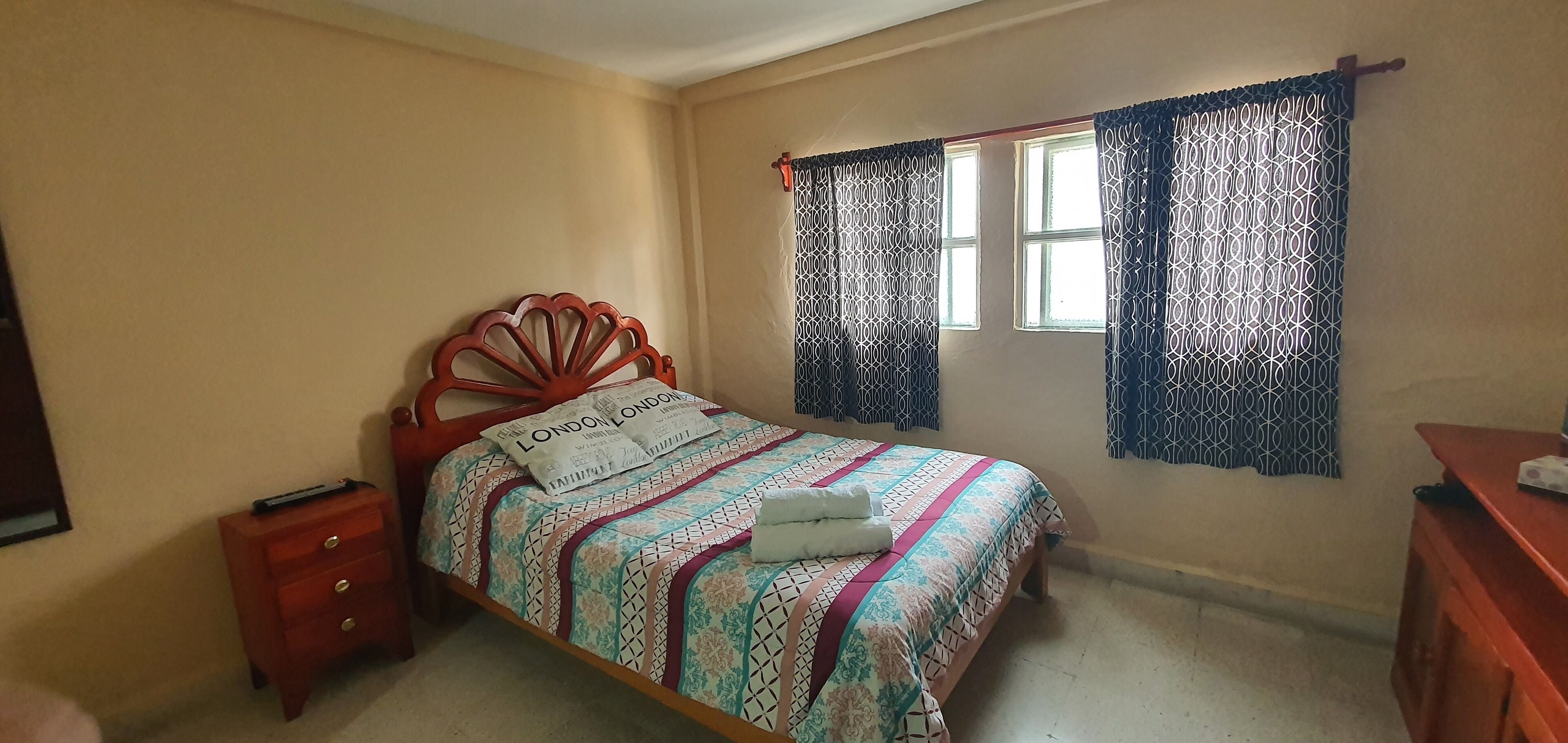 A bedroom with a bed, a nightstand, a wooden dresser, and a window with patterned curtains.