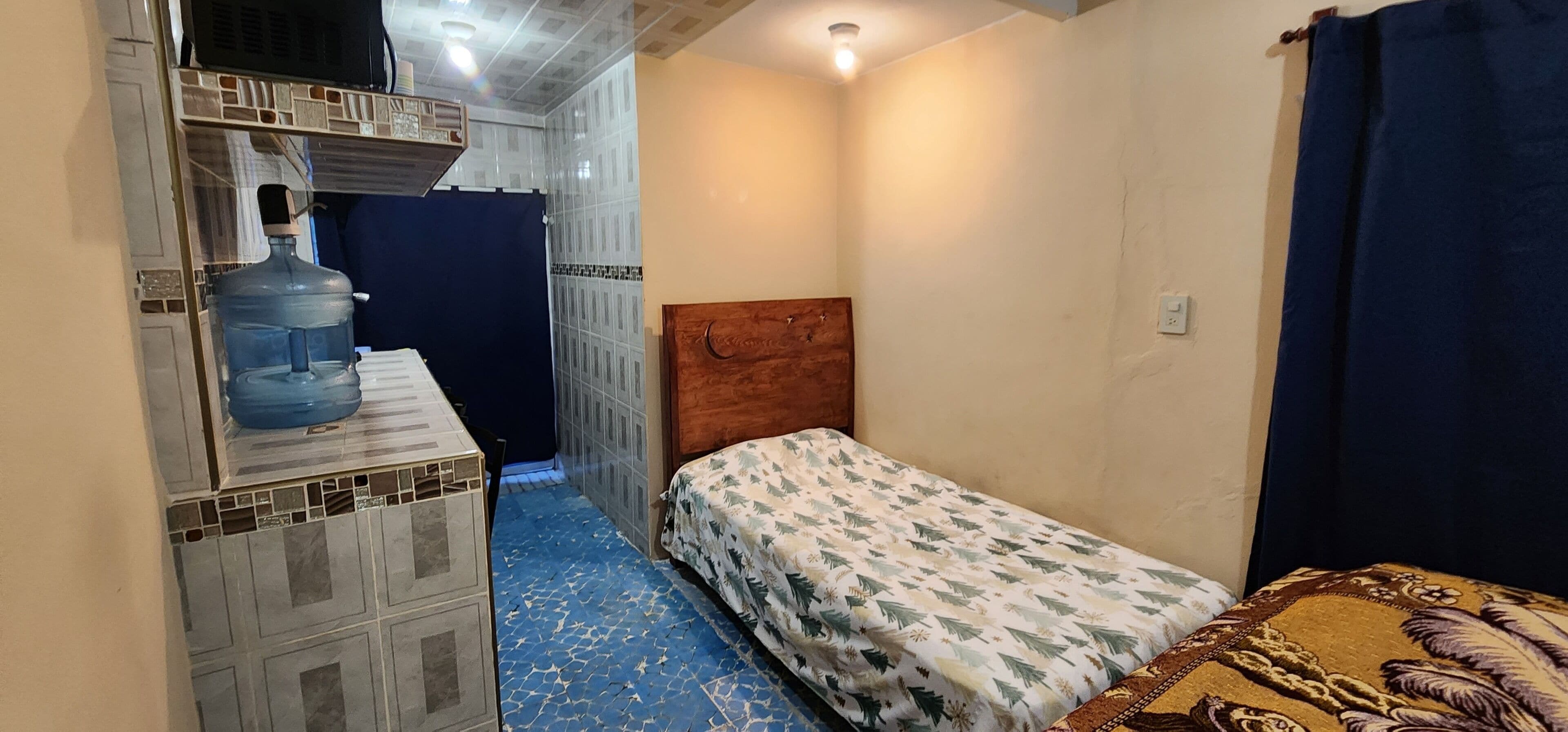Una habitación pequeña y estrecha con una cama, un dispensador de agua y piso de baldosas.