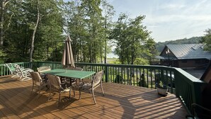 Une terrasse en bois avec une table et des chaises vertes, entourée d’une rambarde verte, d’où l’on a vue sur un lac et des bâtiments.