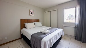 Un lit bien fait, avec du linge de lit blanc et une couverture grise, une tête de lit en bois, un petit tableau encadré accroché au mur et une fenêtre donnant sur des immeubles.