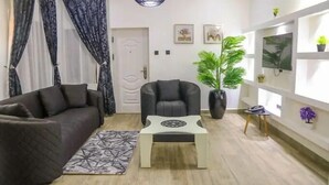 Una sala de estar moderna con un sofá marrón oscuro, un sillón negro, una mesa de centro y un tapete estampado. Se observa una puerta con un picaporte decorativo, una planta en maceta y un televisor montado en la pared.