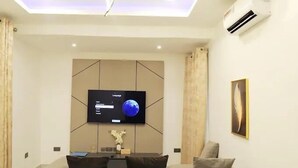 Una sala moderna con un televisor de pantalla plana montado en un panel de pared geométrico, un sofá con cojines y un equipo de aire acondicionado en la pared.