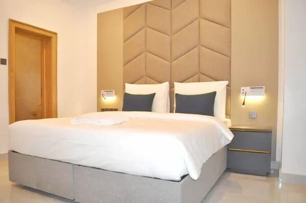 A modern hotel room with a large bed, a tufted headboard, bedside lamps, and a wooden door.