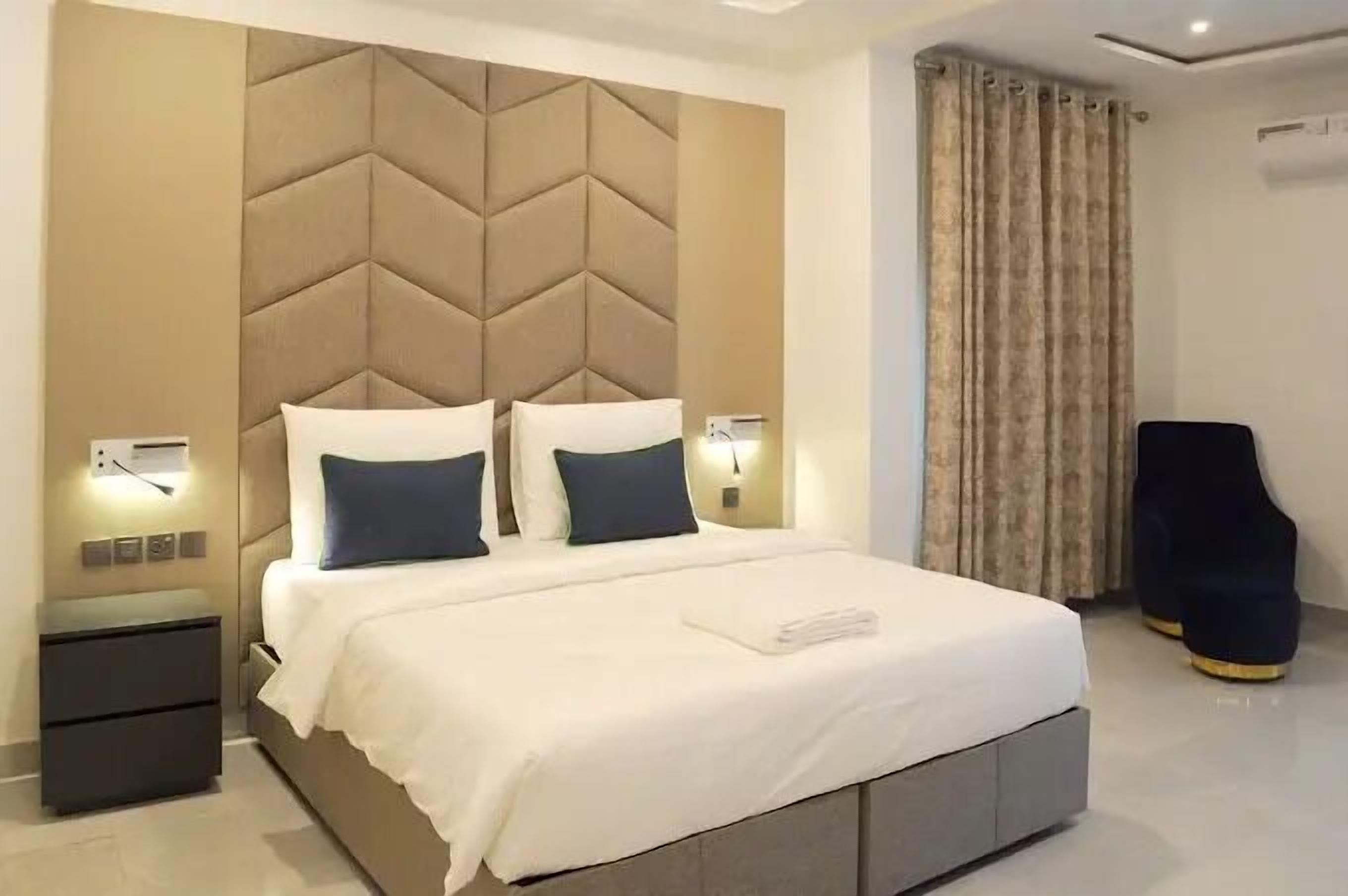Una habitación de hotel moderna con una cama grande, un cabecero con diseño geométrico, lámparas de noche, una mesita de noche y un sillón oscuro.