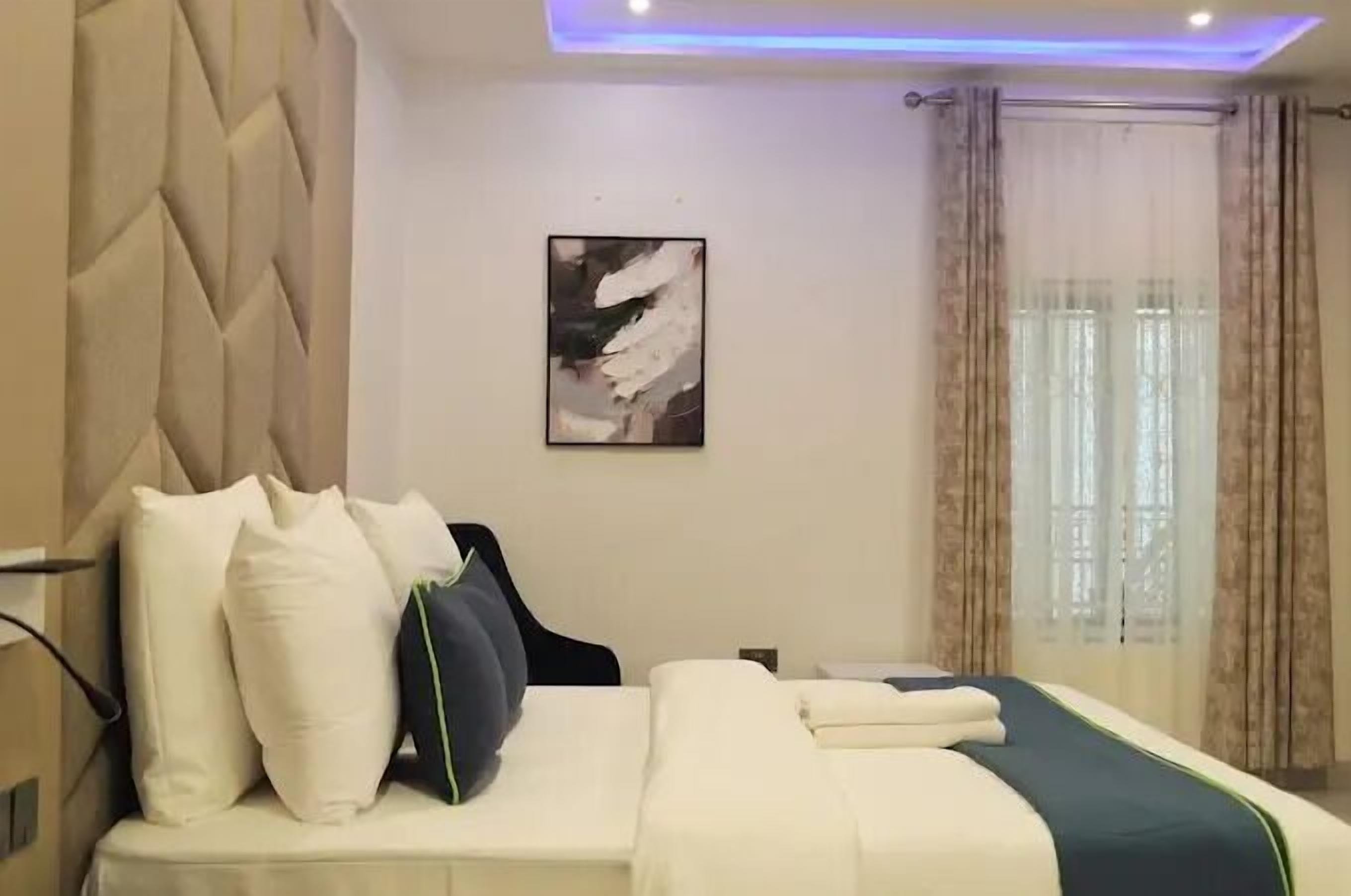 A hotel room with a bed, pillows, a headboard, a wall-mounted artwork, and a window with curtains.