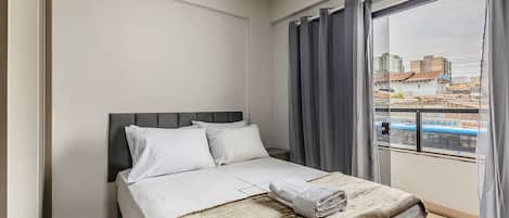 A hotel room with a bed, a window with curtains, and a view of buildings outside.