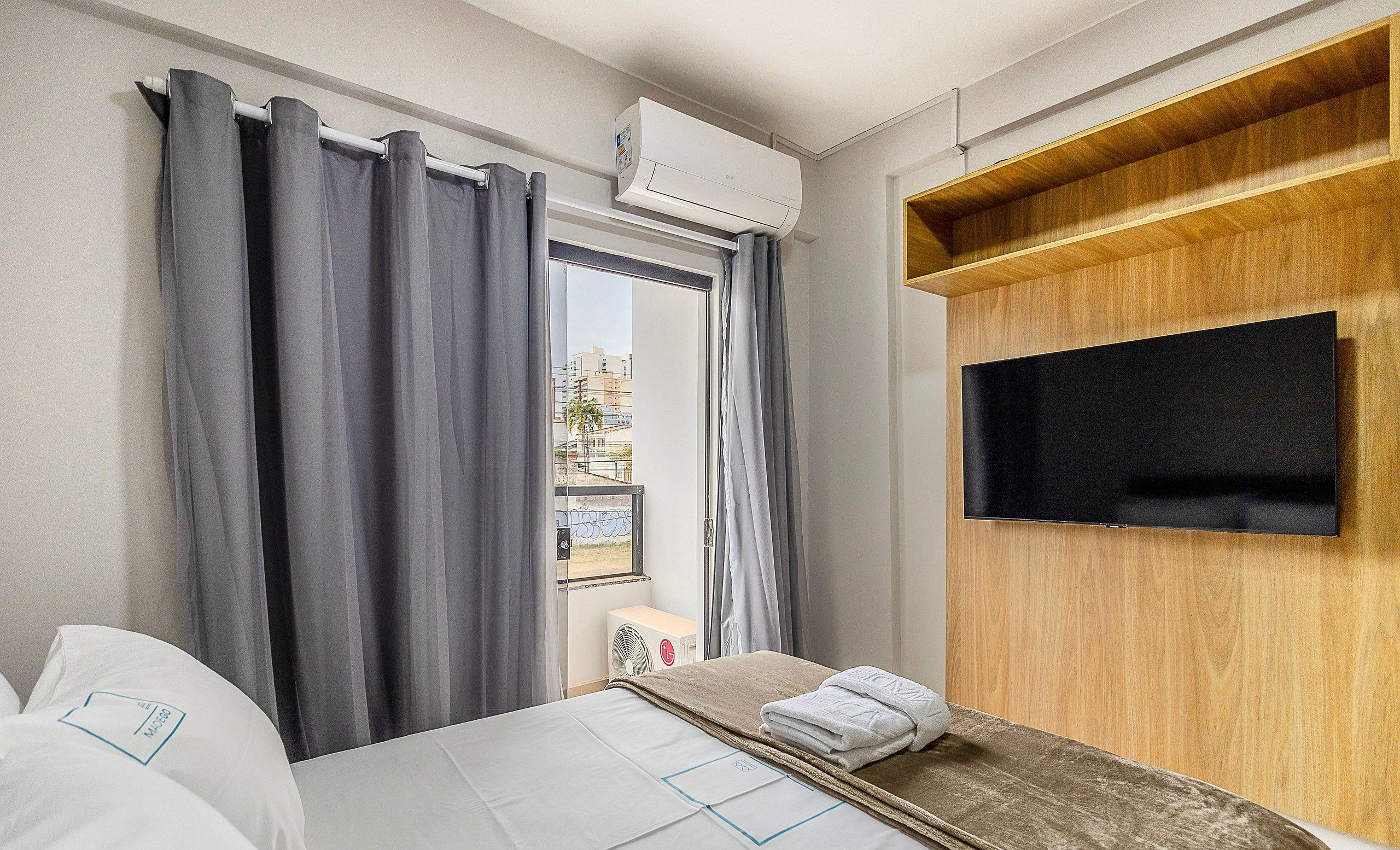Ein Hotelzimmer mit einem Bett, einem an einer Holzplatte montierten Fernseher und einem Fenster mit Vorhängen.