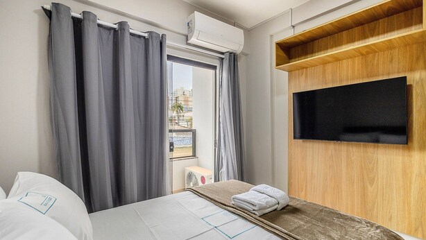 A hotel room with a bed, a TV mounted on a wooden panel, and a window with curtains. - Charming Studio Taguatinga Norte - Rjt0104 (Taguatinga)