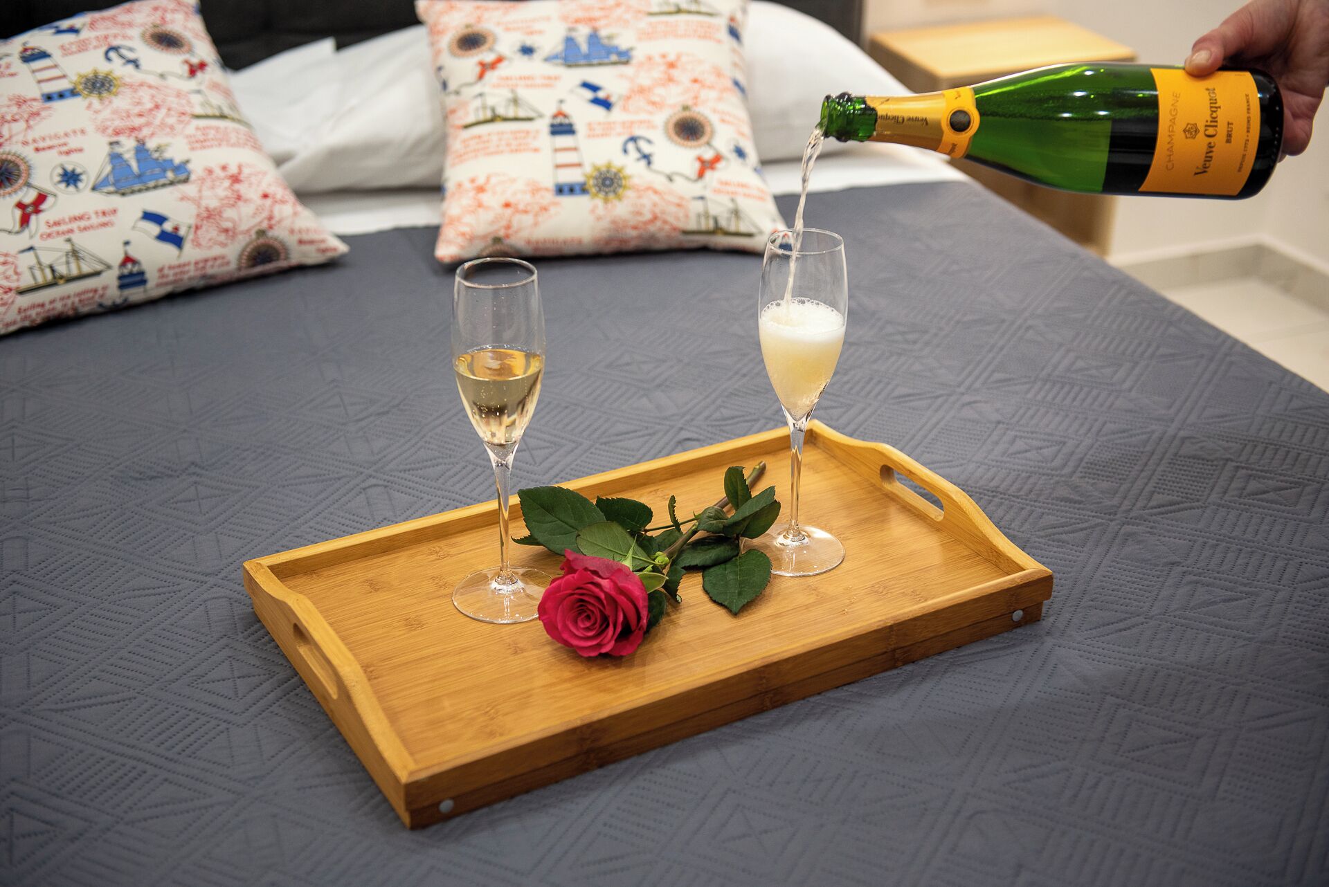 A wooden tray with two champagne glasses and a rose on a bed with patterned pillows.