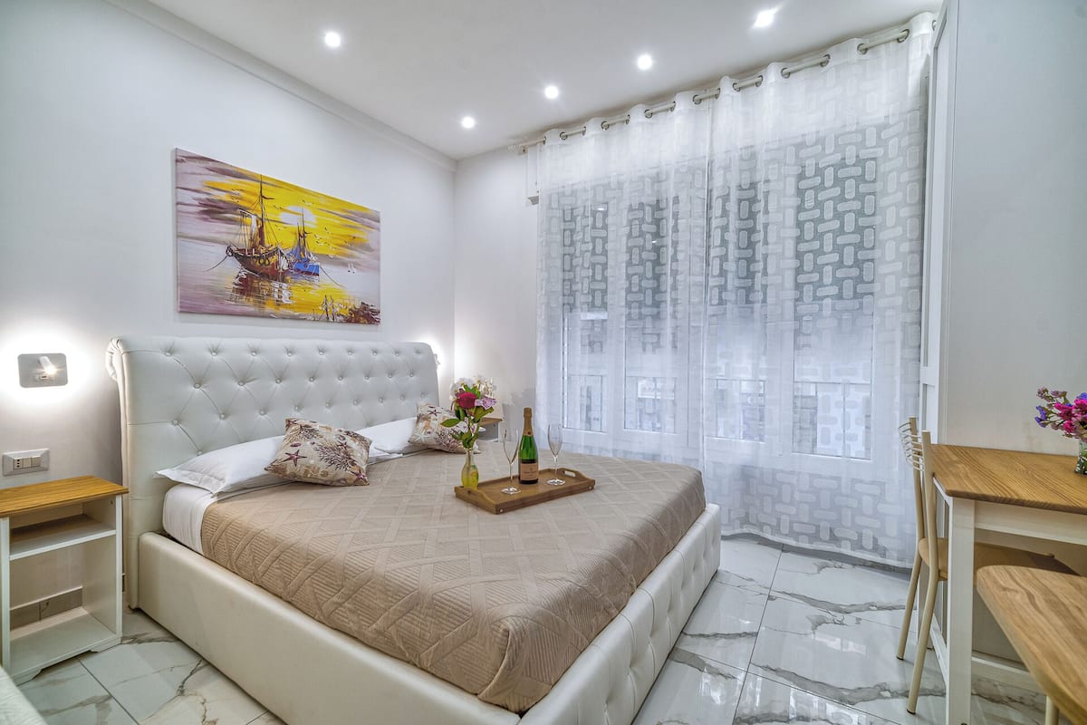 Un dormitorio con una cama de cabecera capitonada, una mesita de noche y una ventana con cortinas estampadas, además de un cuadro en la pared.