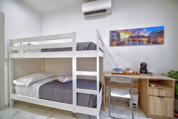 Una cama litera con escritorio y silla, un pequeño cubo de basura y un cuadro de un paisaje urbano.