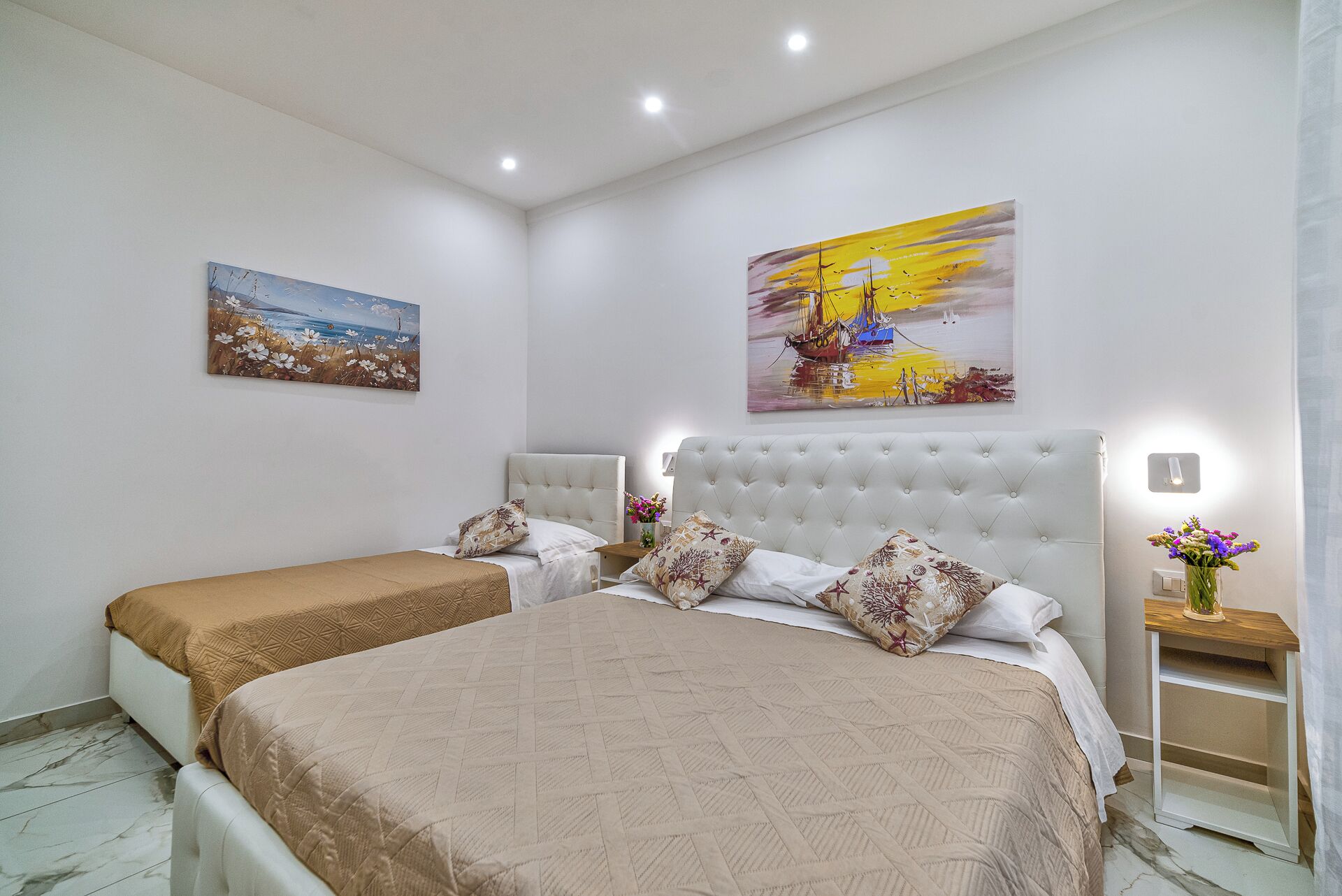 A bedroom with two beds, a large painting, and a nightstand with a vase of flowers.