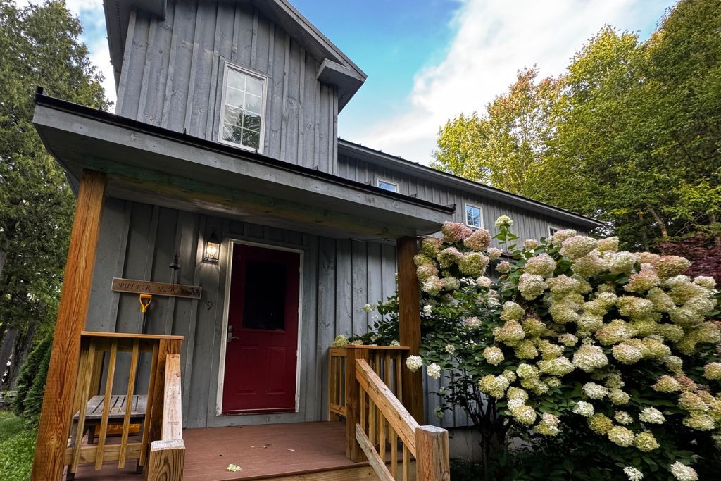 Une maison en bois avec une porte rouge, un porche avec un banc et un grand arbuste d’hortensia.