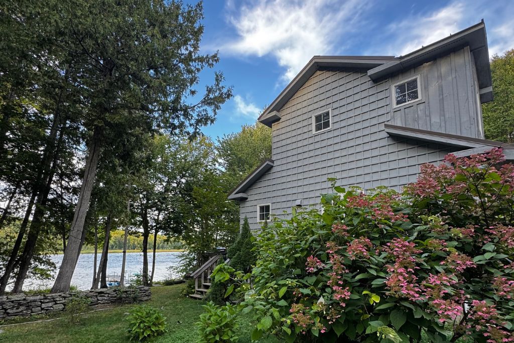 A two-story house with a stone wall and a wooden deck, surrounded by lush greenery and a lake.