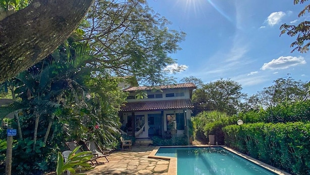 Une piscine dotée d’une terrasse en pierre, entourée d’une végétation luxuriante, avec une maison en arrière-plan.