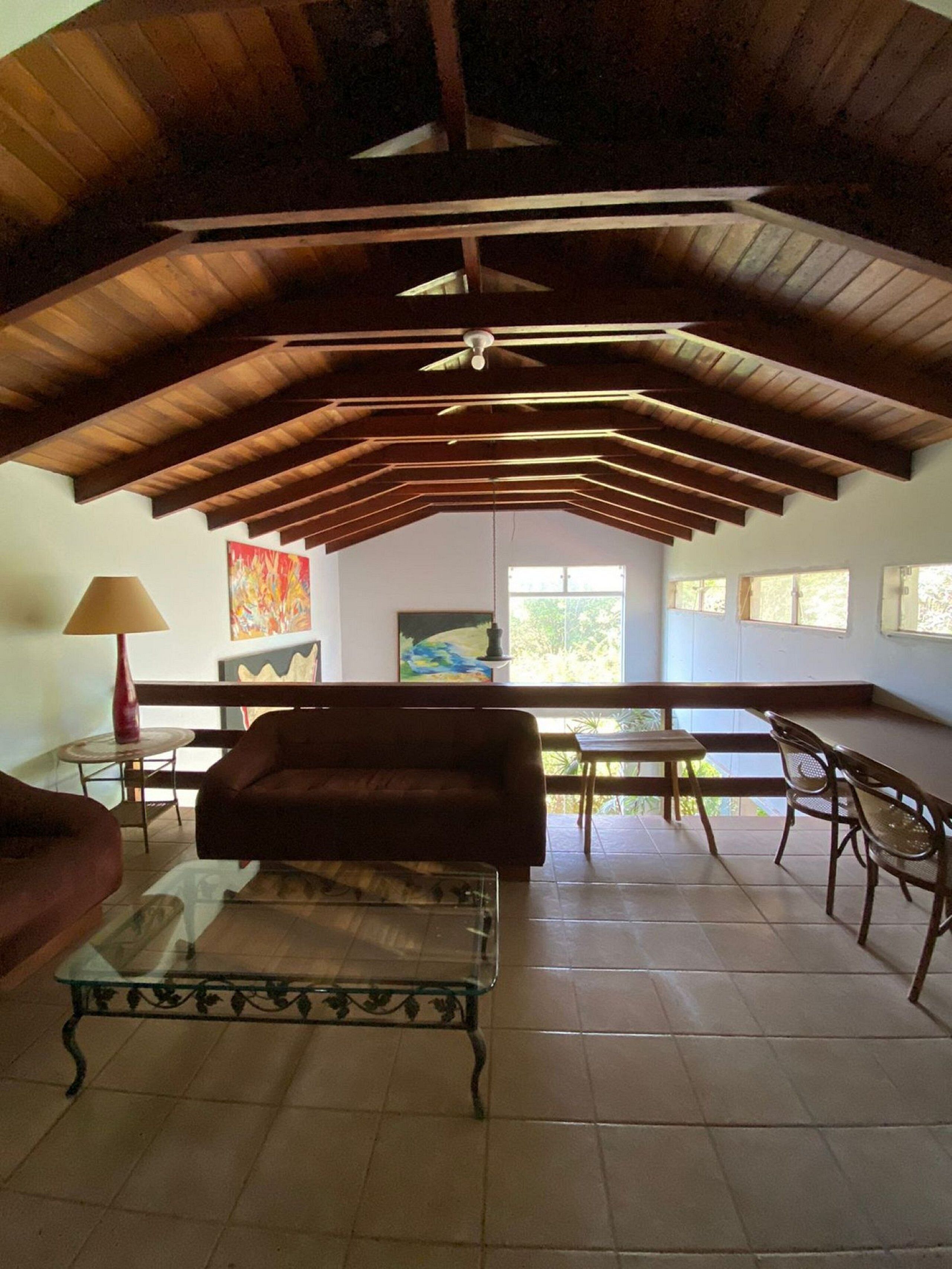 Uma sala de estar ampla com teto alto, vigas de madeira, mesa de centro de vidro, sofá e um espaço de jantar com cadeiras e mesa.