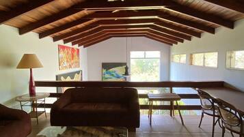 Uma sala de estar ampla com teto alto, vigas de madeira, mesa de centro de vidro, sofá e um espaço de jantar com cadeiras e mesa.