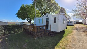 A small, single-story mobile home with a wooden deck and a door, surrounded by other similar homes and trees.