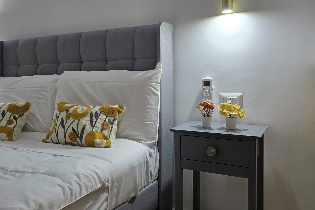 A bedroom with a grey upholstered bed, white bedding, and decorative pillows. There is a small bedside table with a lamp and a potted plant.