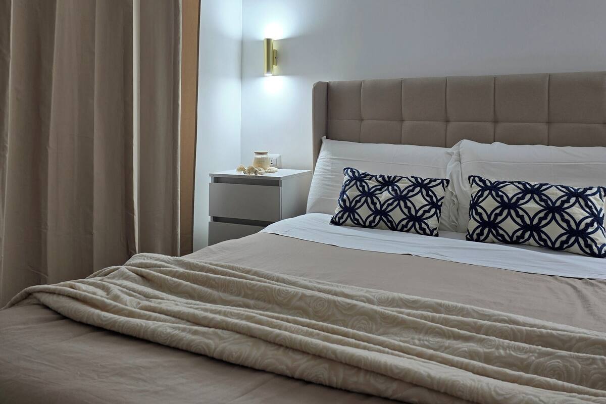 A neatly made bed with a beige comforter and pillows, a bedside table with a lamp, and a window with curtains.