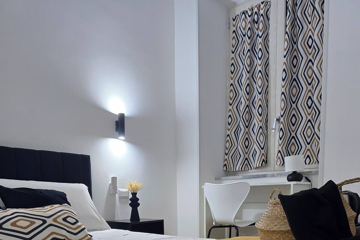 A neatly made bed with a yellow throw blanket, a bedside table with a vase of flowers, and a chair with a woven bag.