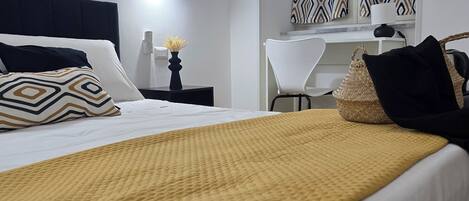 A neatly made bed with a yellow throw blanket, a bedside table with a vase of flowers, and a chair with a woven bag.