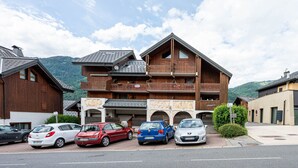 Un immeuble en bois à plusieurs étages, doté de balcons, avec des voitures garées devant et un paysage montagneux en arrière-plan.