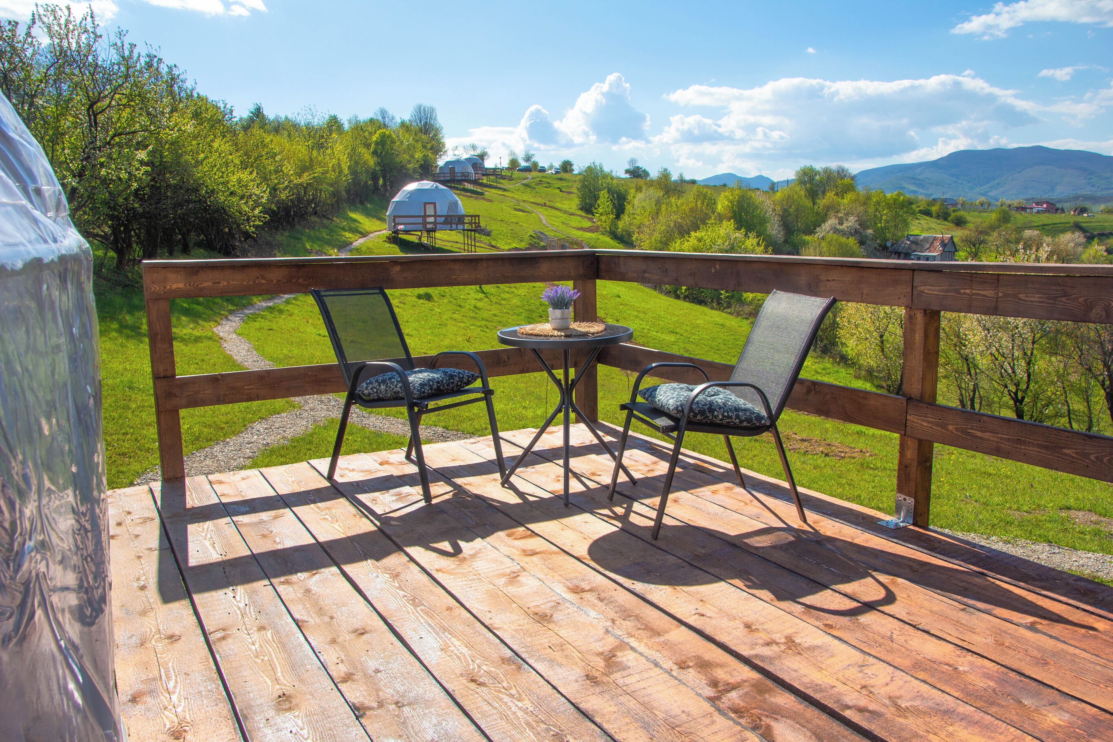 A wooden deck with two chairs and a small table, overlooking a grassy landscape with a dome-shaped structure in the distance.