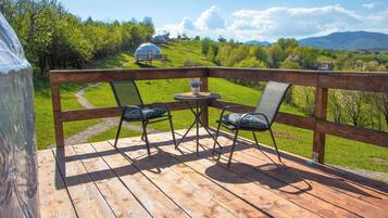 Une terrasse en bois avec deux chaises et une petite table, d’où l’on peut admirer un paysage verdoyant avec, au loin, une structure en forme de dôme.