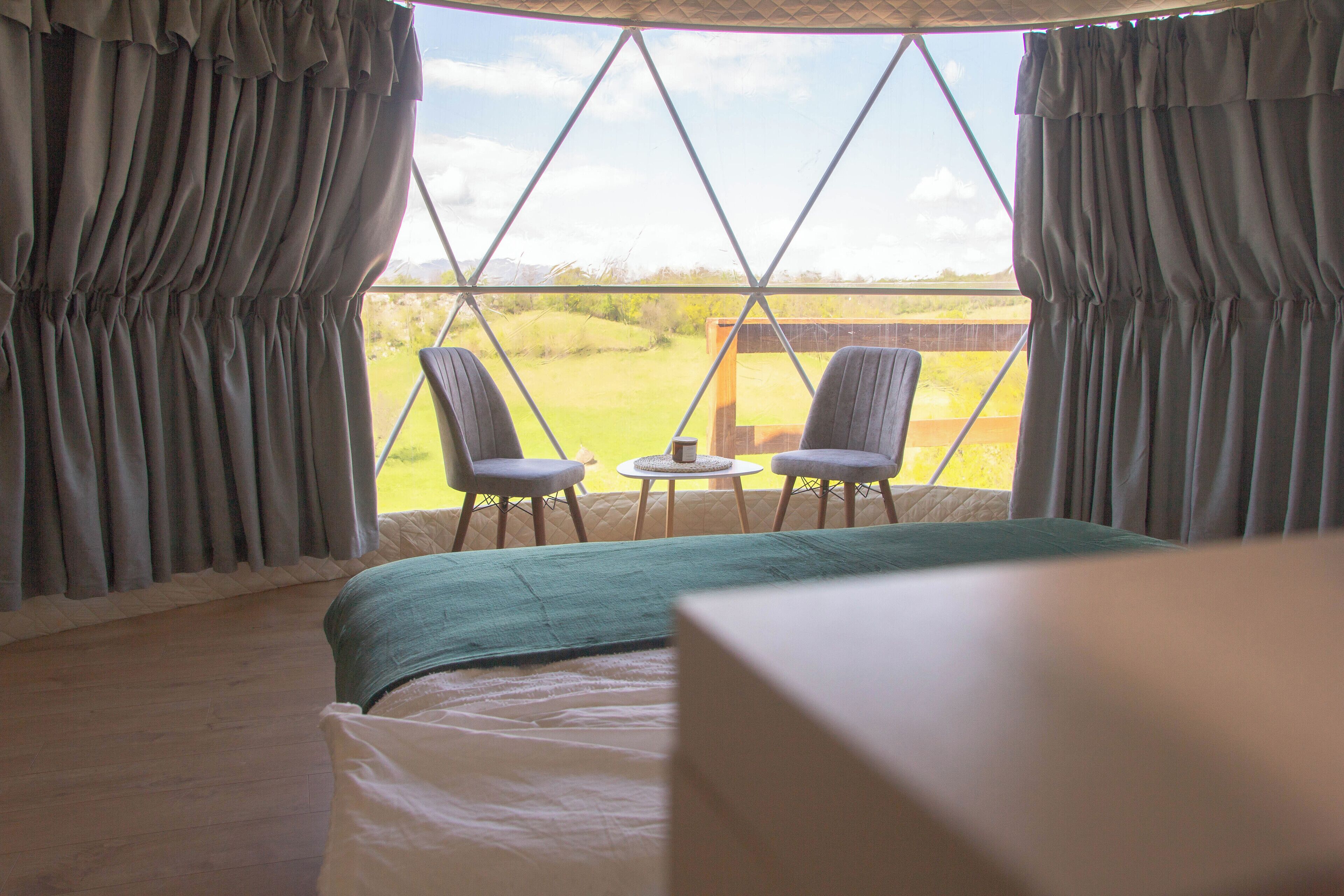 A bedroom with a large window, curtains, a bed, and two chairs facing the window.