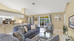 A modern living room with a grey sofa set, a glass coffee table, and a kitchen area with white cabinets and a microwave.