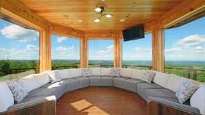 Eine Holzdar Terrasse mit einer gebogenen Sitzgruppe, großen Fenstern und einem Flachbildfernseher.